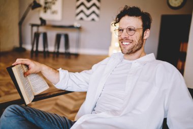 Beyaz tişörtlü, beyaz tişörtlü, sandalyede oturan ve kitap okuyan mutlu, sakallı bir erkek. 