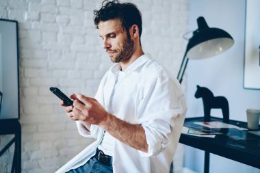 Beyaz tişörtlü ve beyaz tişörtlü endişeli bir erkek akıllı telefona mesaj atıyor. Yaratıcı iş yerinde lamba ve at figürüyle masaya yaslanıyor.