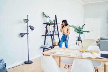 Şık, ferah bir ev ofisinde rafın yanında dururken gülümseyerek ve telefona cevap vererek mutlu siyah kadın.