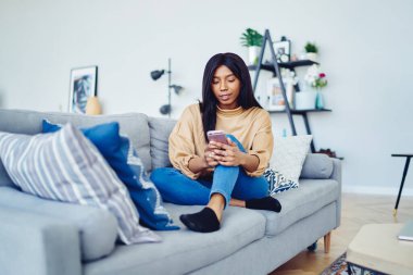 Koyu tenli bir kadın hafta sonu boş zamanlarında modern cep telefonu teknolojisiyle çevrimiçi mesajlaşmaktan zevk alıyor. Ev içi rahat, Afro-Amerikalı blogger 20 'ler sosyal kablosuz ağ kuruyor.