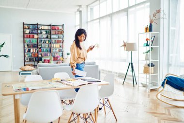 Gündelik giysiler içinde Afrikalı Amerikalı bir kadın. Geniş ofis projesinde çalışırken masasında akıllı telefon düzenleme belgeleriyle.