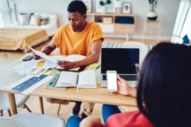 Milenyum blogcusu web sitesini boş akıllı telefondan tarıyor. İnternet reklamları için alanı kopyalıyor. Masada dizüstü bilgisayarlı, koyu tenli adam kağıt belgelerini kontrol ediyor.