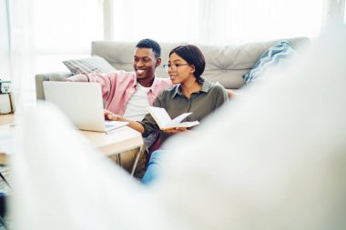 Kanepenin yanında otururken ve serbest proje üzerinde çalışırken dizüstü bilgisayarından gülümseyen ve veri okuyan neşeli siyahi adam ve kadın.