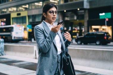 Yuvarlak gözlüklü, resmi takım elbiseli, akıllı telefonlu ve telefonla konuşan saygın kadın girişimcinin yan görüntüsü.