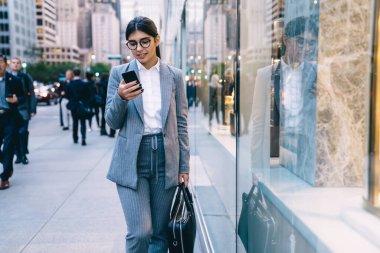 Resmi kıyafetli, gözlüklü, siyah saçlı, siyah çantalı, genç ve güzel bir çalışan. Arka planda yürürken siyah çantalı. 