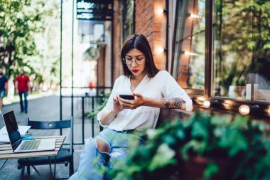 Milenyum kadın blogcusu optik gözlüklerle internet istatistiklerini kontrol ediyor açık hava kafeteryasında açık internet bağlantısını kullanıyor, cep telefonu aygıtında interneti tarayan genç serbest yazar