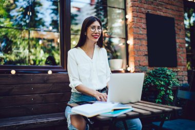 Dürbünlü ve günlük kıyafetlerle gezen mutlu bir kadın. Bilgisayar kullanıyor. Kafenin önündeki bankta oturmuş kameraya bakıyor.