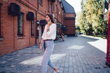 Gözlüklü, beyaz gömlekli, dar kafalı, sokakta yürüyen, elinde klasörlerle cep telefonuyla konuşan genç bir bayan.