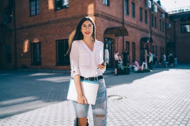 İçerik gözlüklü, uzun saçlı, beyaz gömlekli, kot pantolonlu, elinde not defterleri ve akıllı telefonlu bir Asyalı kadın gündüz vakti sokakta durup kameraya bakıyor.