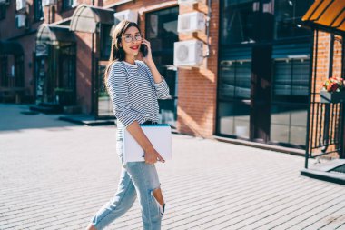 Bilgisayarlı, not defterli, şık giyinmiş, sokakta yürüyen ve cep telefonuyla konuşan, kameraya bakan pozitif içerikli arkadaş canlısı kadın.