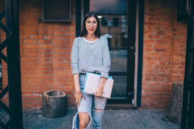 İspanyol kadın, optik gözlüklü, göz koruması için şehir sokağındaki kameraya bakıyor. Modern dizüstü bilgisayar ve eğitim ekipmanlarıyla dışarıda poz veren yetenekli bir kadın.