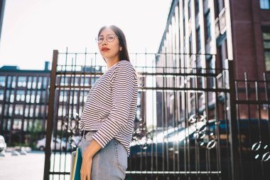 Çağdaş siyah saçlı, çizgili günlük gömlekli ve kırmızı rujlu hippi bir kadının şehir sokaklarındaki kameraya bakışıyla. 