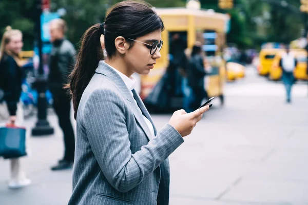 金融街の企業ビルへの途中で4Gのインターネットを使用して携帯電話デバイスの通知を更新する眼鏡でラテン系の女性の側面図 ヒスパニック系弁護士は情報を送信します — ストック写真