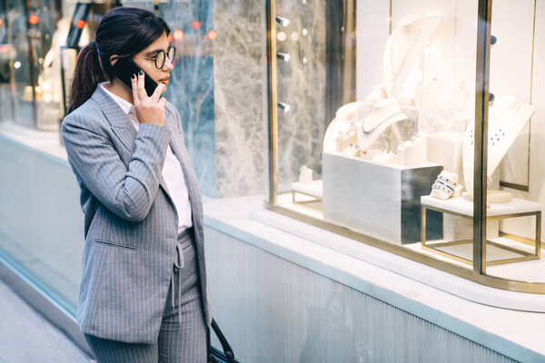 Confident worker female in formal outfit and eyeglasses and with black hair talking on mobile while walking on street on background of blurry people looking at shop 