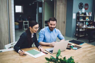 Mavi gömlekli, kolları sıvalı, gülümseyen bir erkek. İş yerinde dizüstü bilgisayar kullanmak isteyen koyu renk saçlı bir kadının yanında oturuyor.