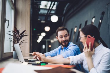 Telefondan konuşan, ekranda kalemle işaret eden neşeli, yetişkin bir çalışanın yan görüntüsü. Aynı zamanda bilgisayarla gülümseyen bir iş arkadaşı ve bulanık bir arka planda modern ofiste birbirlerine bakıyorlar.