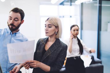 Elinde grafik ve sakallı adam olan sarışın kadın yeni iş stratejisi hakkında beyin fırtınası yaparken arka planda gözlüklü siyah saçlı kadın telefonla konuşuyor.