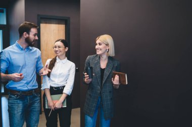 Kahvelerini yudumlayan neşeli genç profesyoneller ellerinde kağıtlarla geziyorlar ve kahkahalarla gülüyorlar ofiste çalışma süreçlerini tartışıyorlar kahverengi duvarlarla. 