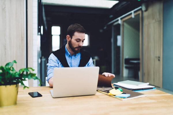 Focused handsome business man wearing blue shirt holding and reading notepad while sitting at desk with laptop in office room