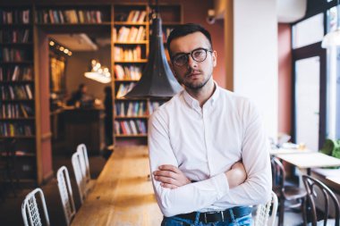 Tişörtlü, gözlüklü, çapraz kollu, hippi kahve dükkanında masaya yaslanmış kameraya bakan düşünceli, modern bir genç.