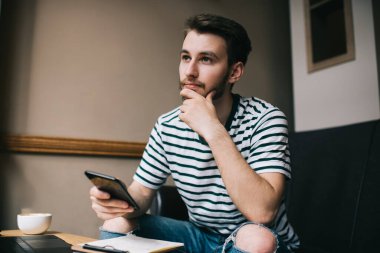 Akıllı bir adam cep telefonu sörfü yapıyor. Masada dizüstü bilgisayarla oturuyor ve koyu ışıklı bir fincan kahve içiyor. 