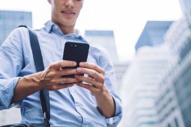 Mavi gömlekli, çantalı, gülümseyen bir adamın modern şehirde gökdelenleri olan bir caddede durup akıllı telefon izlemesi.