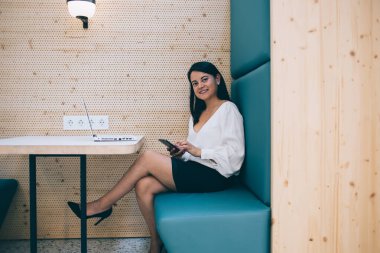 Barcelona Kafe 'de cep telefonu ve dizüstü bilgisayarda sörf yaparken bacakları çapraz oturan ve kameraya bakan resmi giysili siyahi bayanın yan görüntüsü