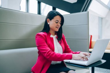 Yoğunlaşmış etnik siyah saçlı bayan parlak iş yerinde masada oturuyor ve iş arkadaşıyla konuşurken bilgisayarını karıştırıyor ve gülümsüyor.