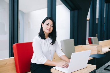 Genç bayan girişimci, modern açık uzay ofisinde otururken laptop kullanıyor ve kameraya bakarken gülümsüyor.