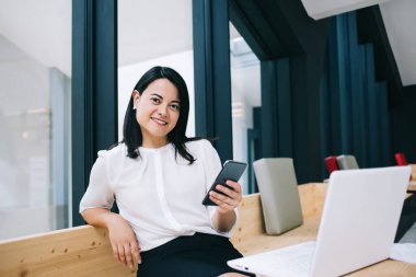 Resmi giyinmiş genç bir kadın, rahat bir şekilde bilgisayarın karşısında oturmuş gülümserken telefon kullanıyor.