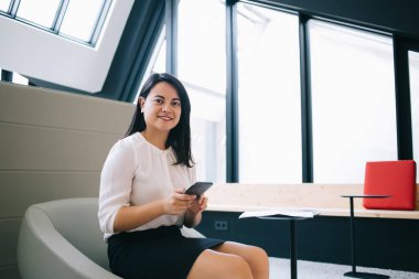 Şık elbiseli neşeli bayan yönetici koltukta oturuyor ve ara verip kameraya bakarken cep telefonunu dinliyor.