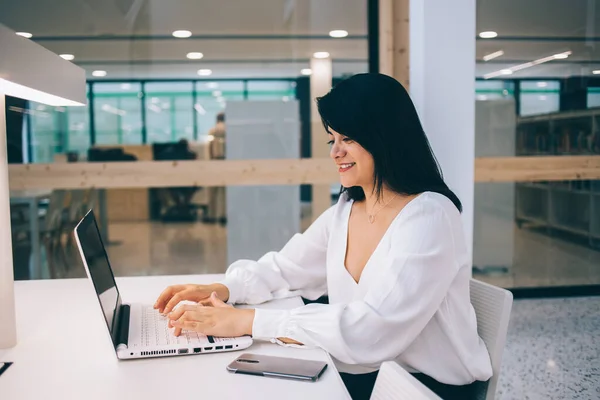 穿着正式服装的年轻女企业家坐在办公桌前 兴高采烈地在笔记本电脑上打字 同时在宽敞而轻盈的办公室里工作的侧视图 — 图库照片