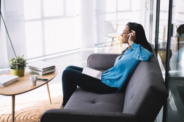 Rahat ve neşeli Afrikalı Amerikalı çalışanın yan görünüşü parlak ofiste koltukta oturmuş tabletle müzik dinlerken gülümsüyor.