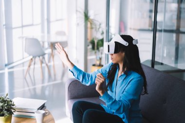 Kanepede oturan, VR kulaklık takan ve sanal gerçeklikle ilgili yeni deneyimler edinen genç bir kadının yan görüntüsü.