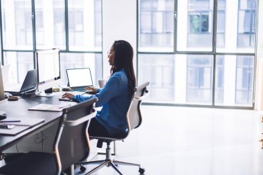 Modern çalışma alanındaki kırtasiyeyle ofis masasında oturan ve bilgisayarda yazı yazan ciddi yoğunlaştırılmış etnik kadın.