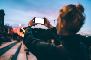 Tanımlanamayan gözlüklü bir kadının arka planda boş akıllı telefon ekranına bakması ve merdivenlerde durup günbatımının fotoğrafını çekmesi.