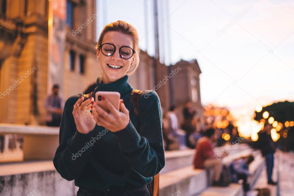 Excited hipster girl rejoicing while using 4g wireless for networking and online communication, cheerful female tourist in classic spectacles blogging creating publication about tourism and trip