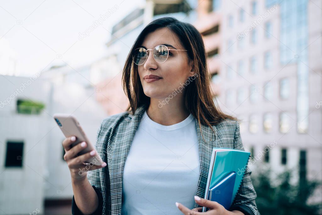 Attractive millennial hipster girl in eyewear using modern smartphone for chatting online standing on urban settings, beautiful caucasian student send text messages on mobile phone using 4G connection