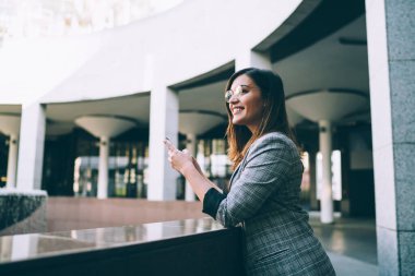 Modern bina lobisinde boş zamanlarında dinlenirken, gözlüklü, akıllı telefonlu neşeli zarif bir kadının düşük açılı yan görüntüsü.