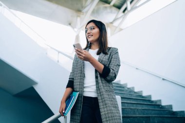 Moda giysileri ve gözlüklü beyaz bir kadın cep telefonunu arıyor merdivenlerde yürüyor, milenyum kadın blogcusu akıllı telefondan 4G bağlantısı ile multimedya paylaşıyor.