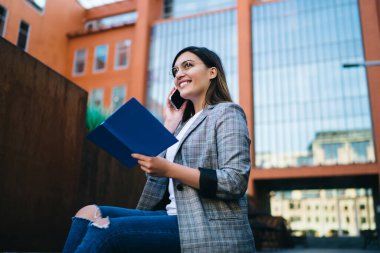 Yanında gözlüklü, sıradan giysileri olan, elinde not defteri olan ve üniversitenin yanındaki merdivenlerde oturan genç ve çekici bir kadının cep telefonuyla konuşması.
