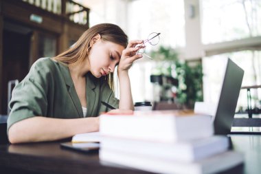 Uzun saçlı, elinde gözlük tutan, gözlerini kapatan, hafif kütüphanedeki laptopta oturan sıkkın ve zeki öğrencinin yan görünüşü.