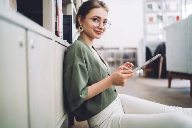 Sıradan giyinmiş, ofis zemininde oturmuş, aletlerle çalışırken kameraya bakan genç bir kadının yan görüntüsü.