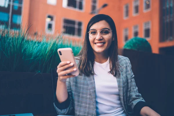 快乐的年轻迷人女性 戴着眼镜 看着相机 一边用智能手机冲浪一边坐在外面的咖啡店里休息 — 图库照片