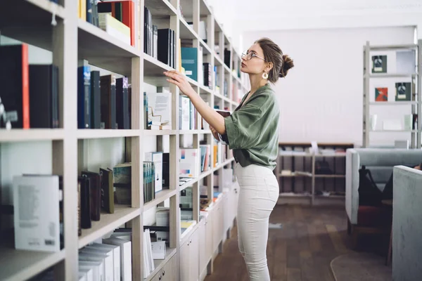 高校图书馆里 穿着眼镜和绿色衬衫 站在书架上看书的时髦女青年的侧视图 — 图库照片