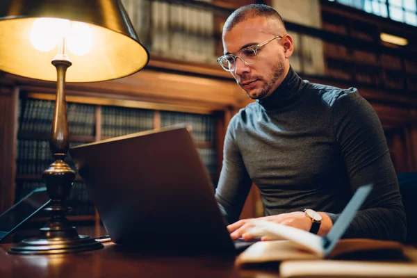 Adult 30 years old male student spending time in university library for learning, intelligent man in optical bifocal spectacles searching scientific information via app on modern laptop computer