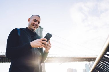 Siyah atletizm takımında formda bir adamın akıllı telefonuyla mesajlaşması ve modern şehir köprüsünün arka planında parlak bir ışık altında gülümsemesi. 