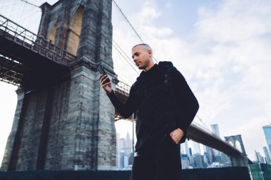 Özenli, kısa saçlı, siyah spor giysili genç adam şehir planında güpegündüz köprüde akıllı telefonları dinliyor.