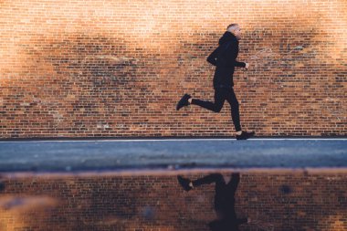 Sağlıklı kısa saçlı, siyah spor giysili ve spor ayakkabılı genç adam dar kaldırımlarda koşuyor.