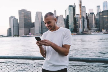 Yakışıklı, kısa saçlı, kulaklıkla müzik dinleyen, akıllı telefon kullanırken gülümseyen ve nehir ve şehrin bulanık arka planına dayanan etnik bir adam.